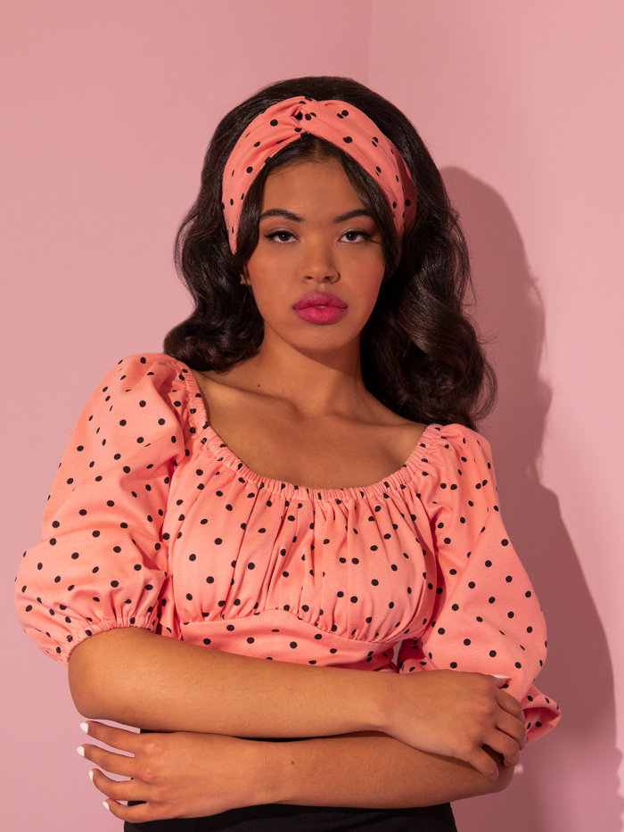 Danelly poses with her arms held across her torso wearing the Vintage Style Knot Headband in Peach Pink Polka Dot and matching vintage inspired top.