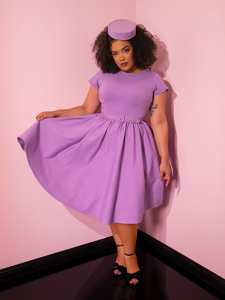 Female model standing in a pink walled showroom wearing the Avon Swing Dress in Lilac and holding up the skirt section to the side to show off the vibrant color.