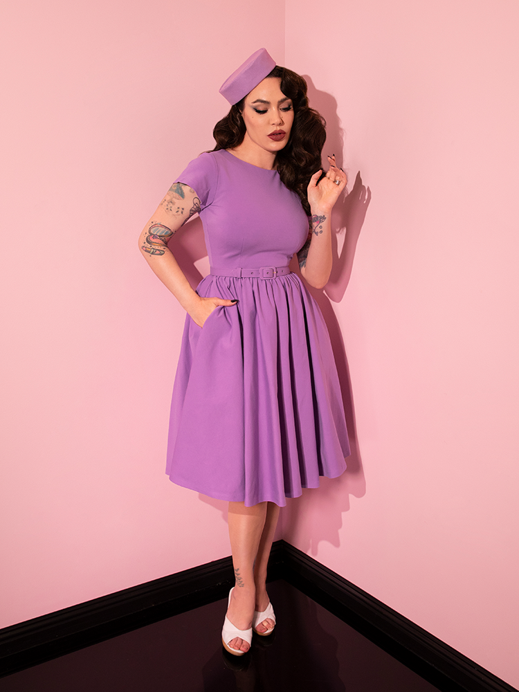 Micheline Pitt posing with one hand in her pocket and the other held up near her face while wearing the Avon Swing Dress in Lilac.