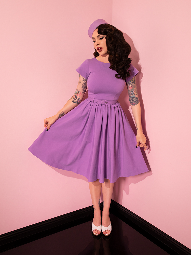 Micheline Pitt wearing a lilac colored retro style dress pulling out the sides to show off the skirt section.