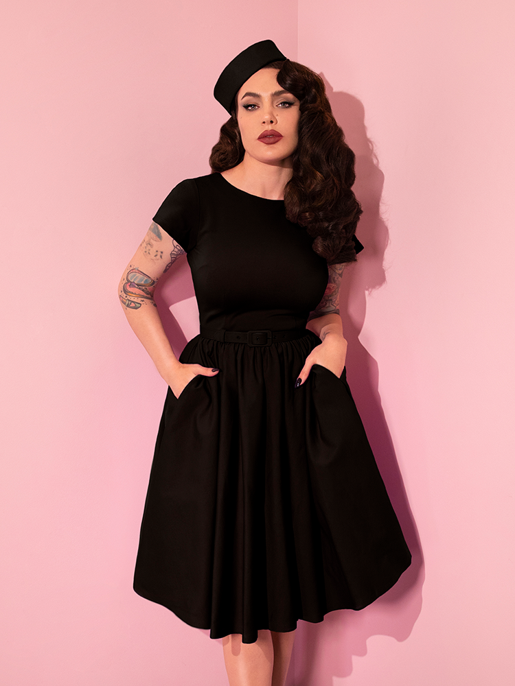 Micheline Pitt wearing the Avon Swing Dress in Black while tucking her hands into the hidden side pockets.