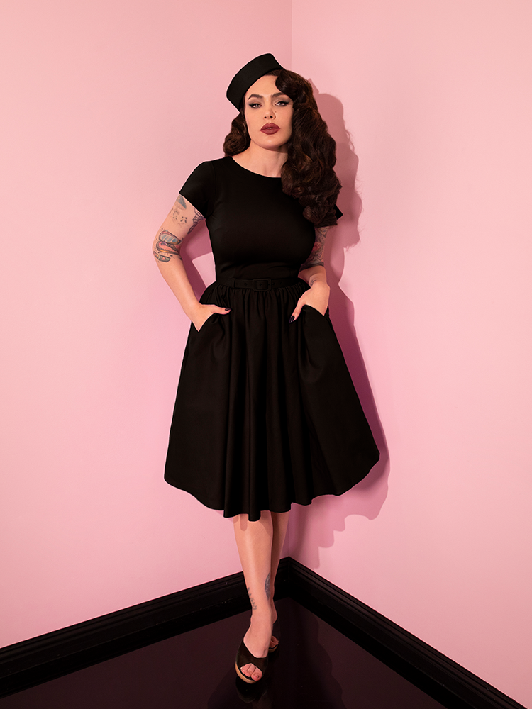 Full length shot of Micheline Pitt posing in the Avon Swing Dress in Black with her hands tucked into the side pockets. Also wearing a complimentary pill box hat. All items available from Vixen Clothing.