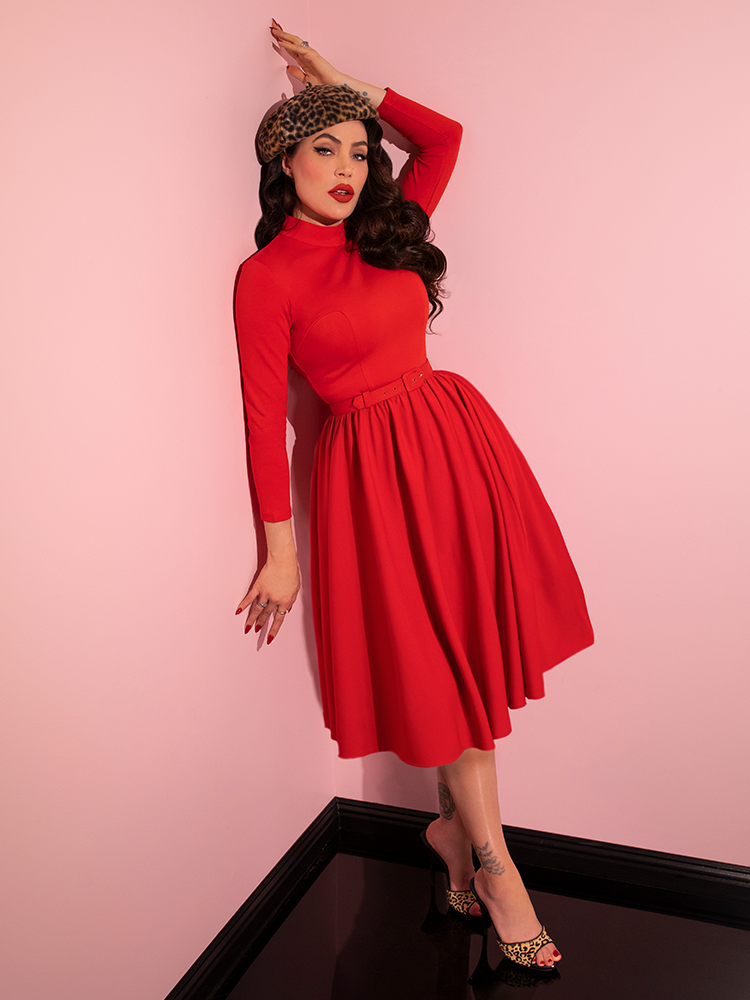 Micheline Pitt leaning against the wall while modeling the Bad Girl Swing Dress in Tomato Red from retro clothing company Vixen Clothing.