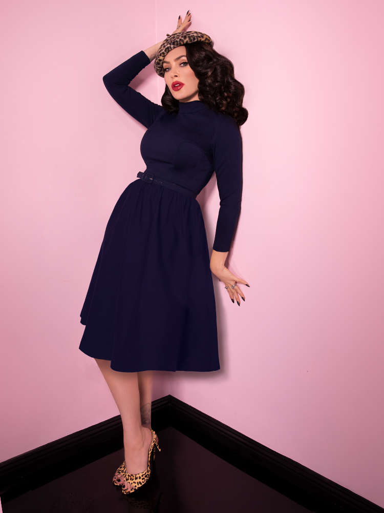 Full body length shot of Micheline Pitt leaning against pink walls while wearing the Bad Girl Swing Dress in Navy