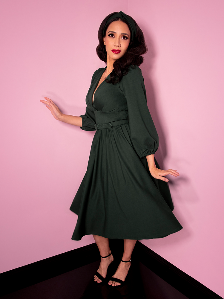 Posing and showing the camera a slight smirk, Milynn Moon is photographed in the Bawdy Swing Dress in Hunter Green.