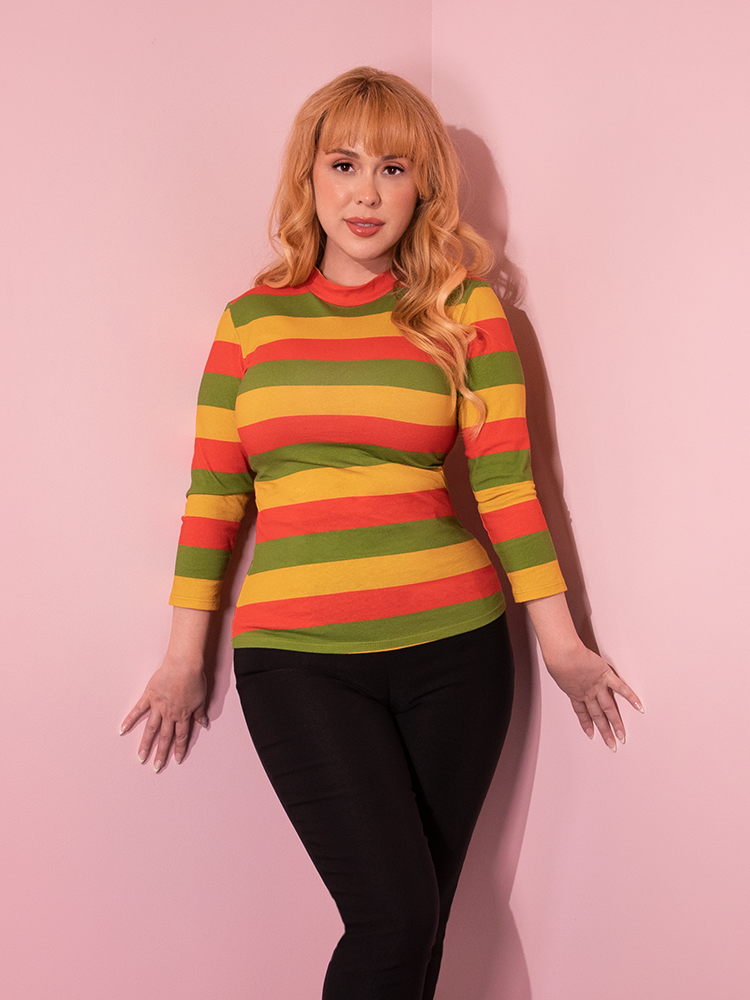 Model posing in the corner of a room wearing the Bad Girl 3/4 Sleeve Top in Orange/Yellow/Avocado Stripes.