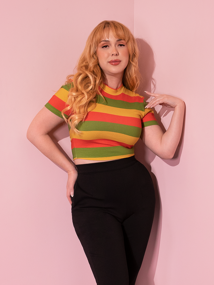 Female model standing a corner wearing the Bad Girl Crop Top in Orange/Yellow/Avocado Stripes.