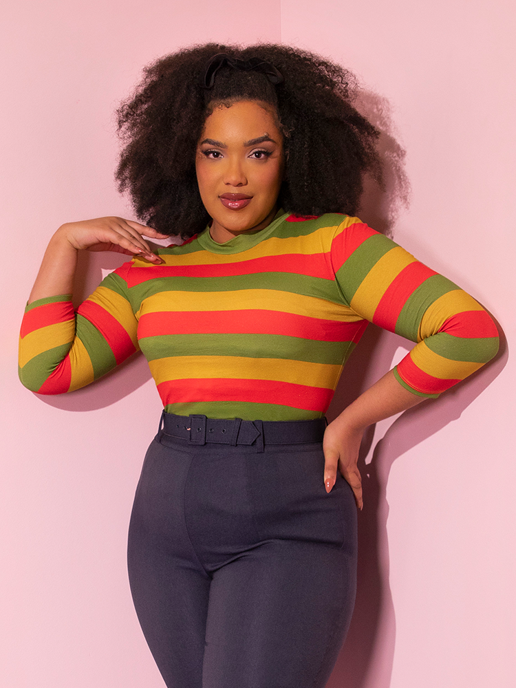 Model posing while wearing the Bad Girl 3/4 Sleeve Top in Orange/Yellow/Avocado Stripes tucked into dark blue form-fitting pants.