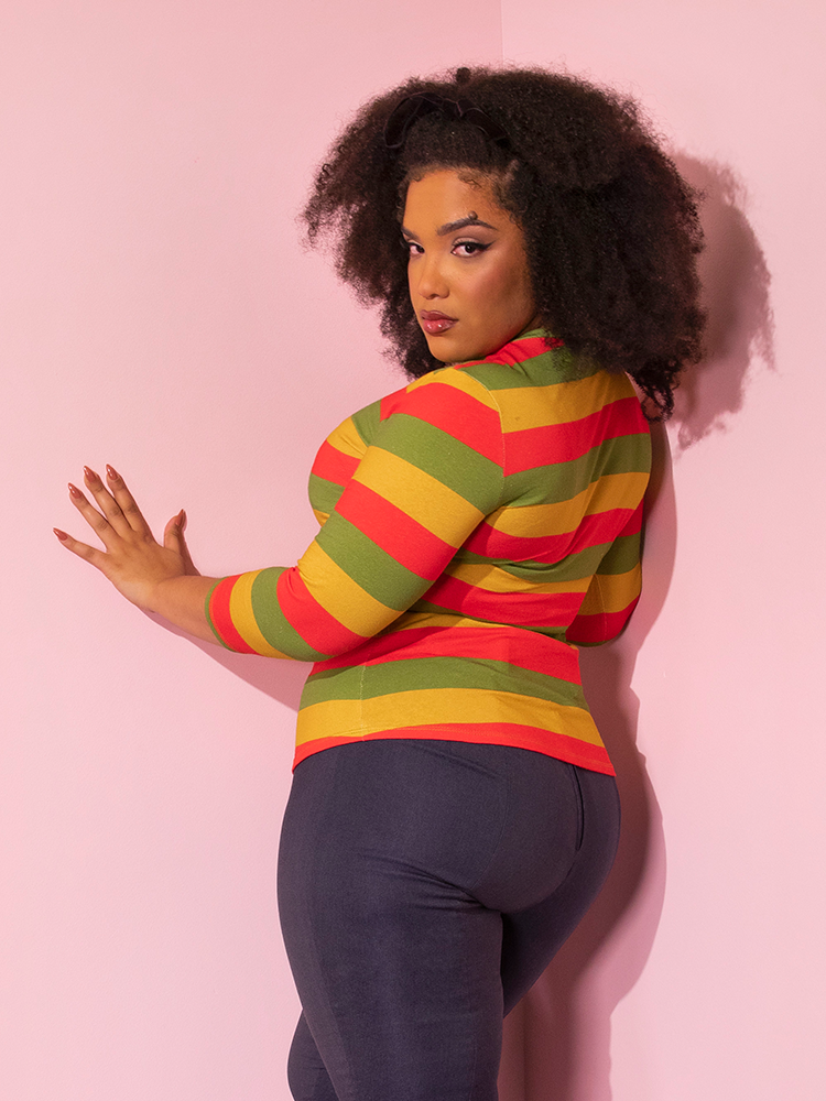 Female model turned away from the camera, but looking back at it while wearing the Bad Girl 3/4 Sleeve Top in Orange/Yellow/Avocado Stripes paired with stretchy denim colored pants.