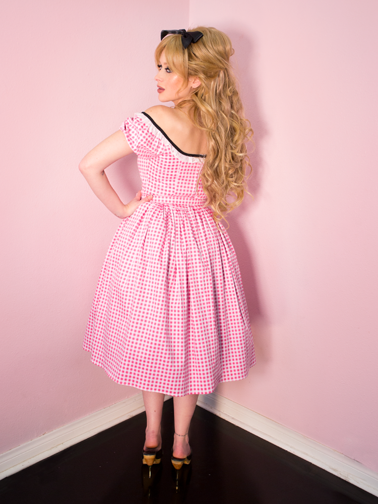 Model turned towards the wall and away from the camera, while wearing a retro style dress in pink gingham print with a partly open back.