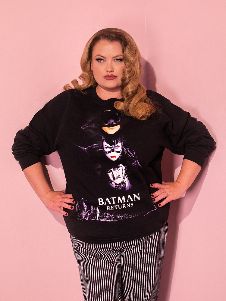 A playful dirty blonde-haired female model exudes charm while posing in the unisex BATMAN RETURNS™ Movie Poster Sweatshirt from the retro clothing brand Vixen Clothing.