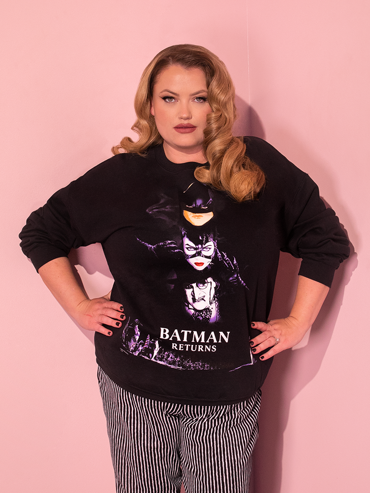 The BATMAN RETURNS™ Movie Poster Sweatshirt (unisex) from Vixen Clothing, a renowned retro brand, is showcased by an attractive dirty blonde-haired model in a playful and charming pose.