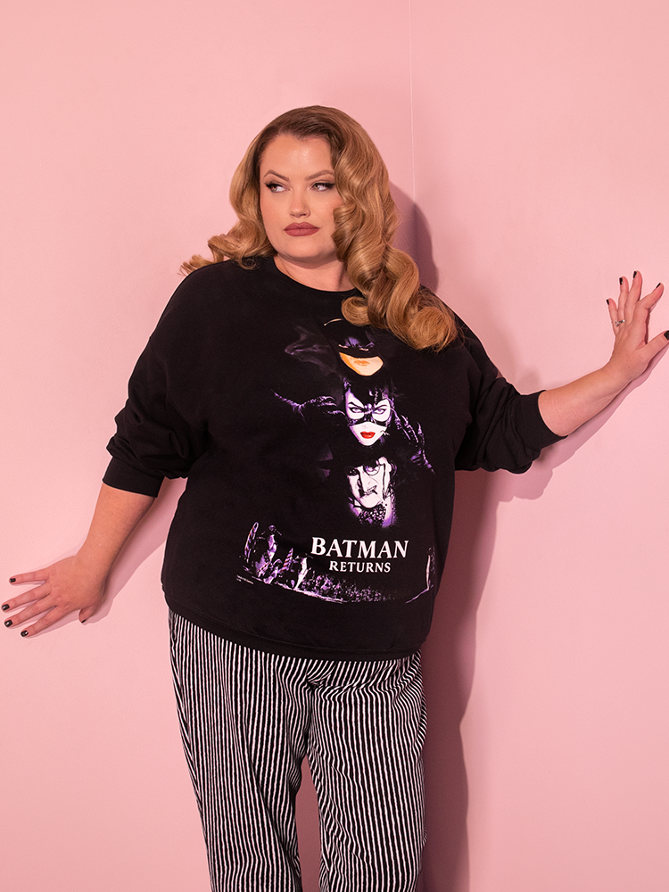The playful pose of an attractive dirty blonde-haired model highlights the retro charm of the BATMAN RETURNS™ Movie Poster Sweatshirt (unisex) from Vixen Clothing.