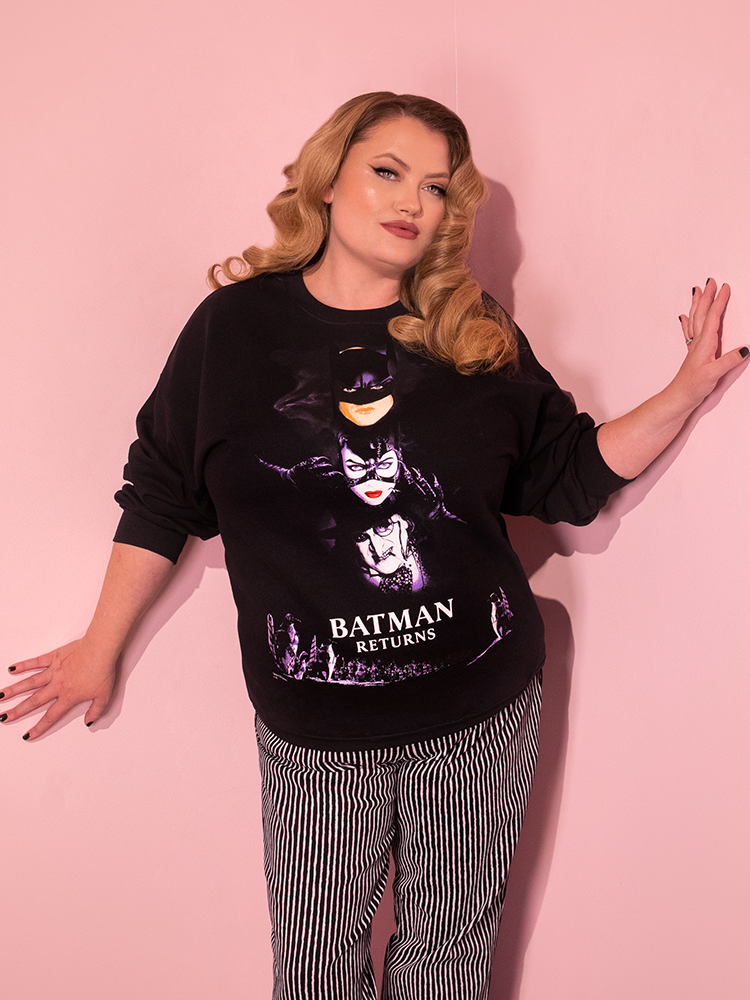 With a playful flair, an attractive dirty blonde-haired model strikes a pose in the BATMAN RETURNS™ Movie Poster Sweatshirt (unisex) from the retro clothing brand Vixen Clothing.