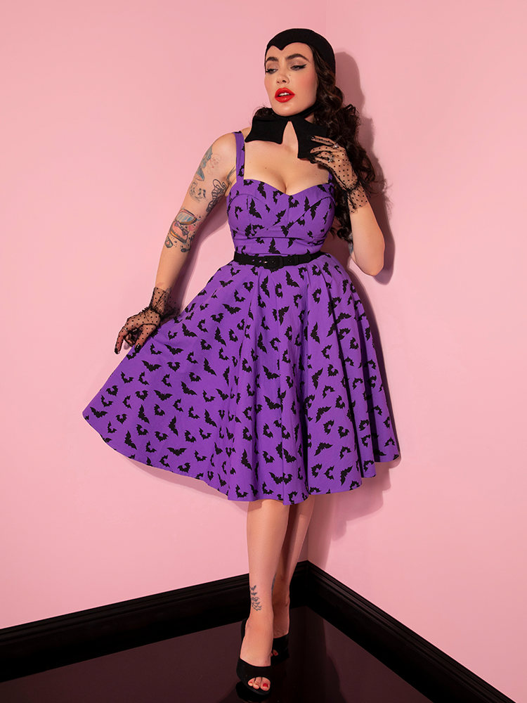 Micheline Pitt standing in the corner of her pink showroom wearing the Maneater Swing Dress in Bat Print from retro dress company Vixen Clothing.