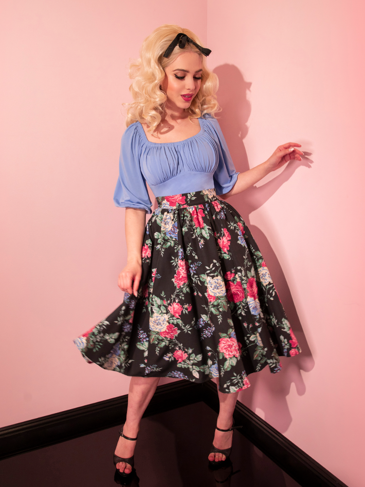 Full length shot of blonde haired model wearing the Vacation Swing Skirt in Black Vintage Rose Print along with a corn blue peasant top.