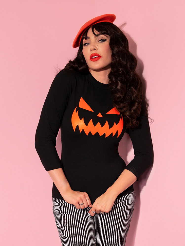 Micheline Pitt looks directly at the camera while gently tugging down on the front of her Pumpkin King 3/4 Sleeve Top in Black and Orange.
