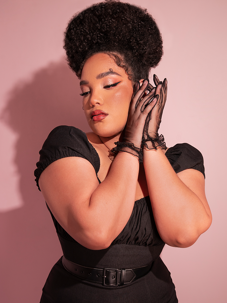 Ashleeta holding both of her hands next to the left side of her head while wearing the Sheer Wristlet Gloves in Black from retro inspired clothing brand Vixen Clothing.