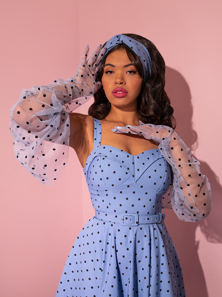 A model posing in the corner of a pink room wearing a corn blue polka dot retro dress paired with Mesh Polka Dot Full Length Gloves in Sunset Blue.