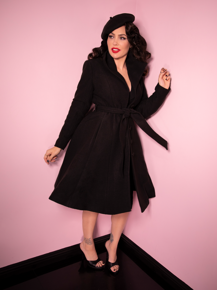 Smiling and looking off-camera, Micheline Pitt channels her best 60s era British spy look with the Starlet Swing Coat in Black with matching accessories.