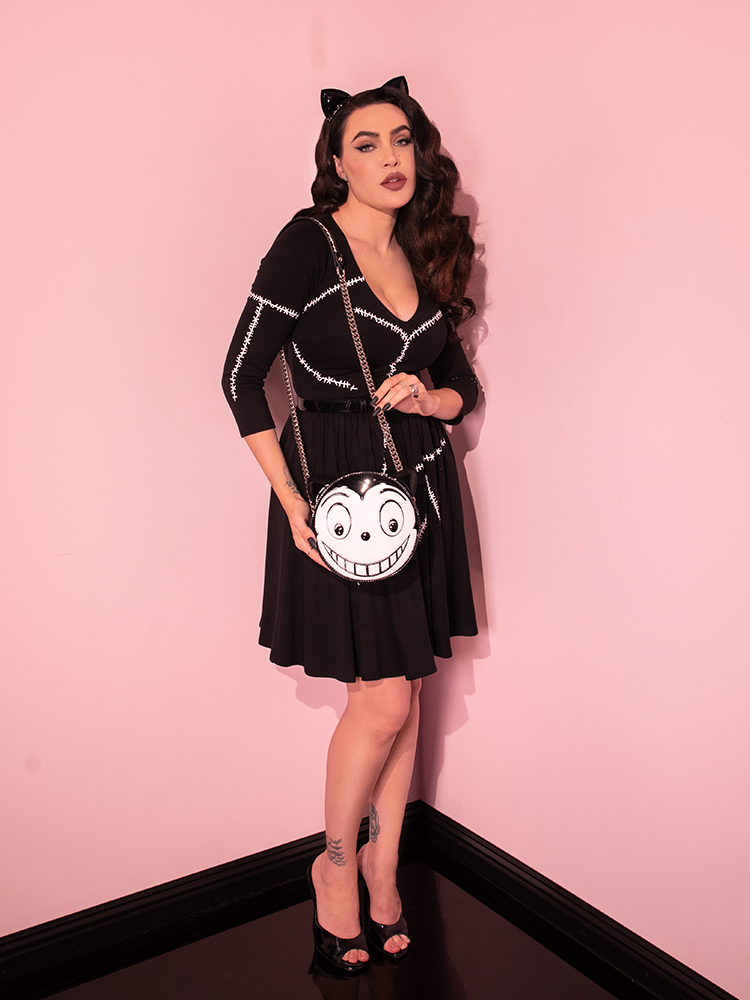 Micheline Pitt posing in the BATMAN RETURNS™ Catwoman Stitch Swing Dress with Catwoman themed purse draped over her right shoulder.
