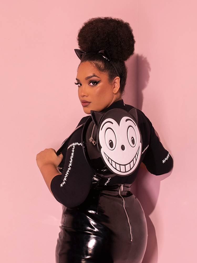 Ashleeta facing away from the camera, but looking over her shoulder, wears the BATMAN RETURNS™ Catwoman Stitches Bad Girl Top With a similarly themed backpack.