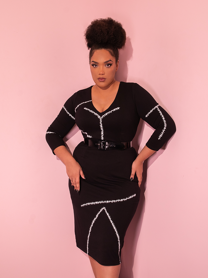 Ashleeta poses in the corner of a pink showroom while wearing the ATMAN RETURNS™ Catwoman Stitch Wiggle Dress from retro style dress brand Vixen Clothing.