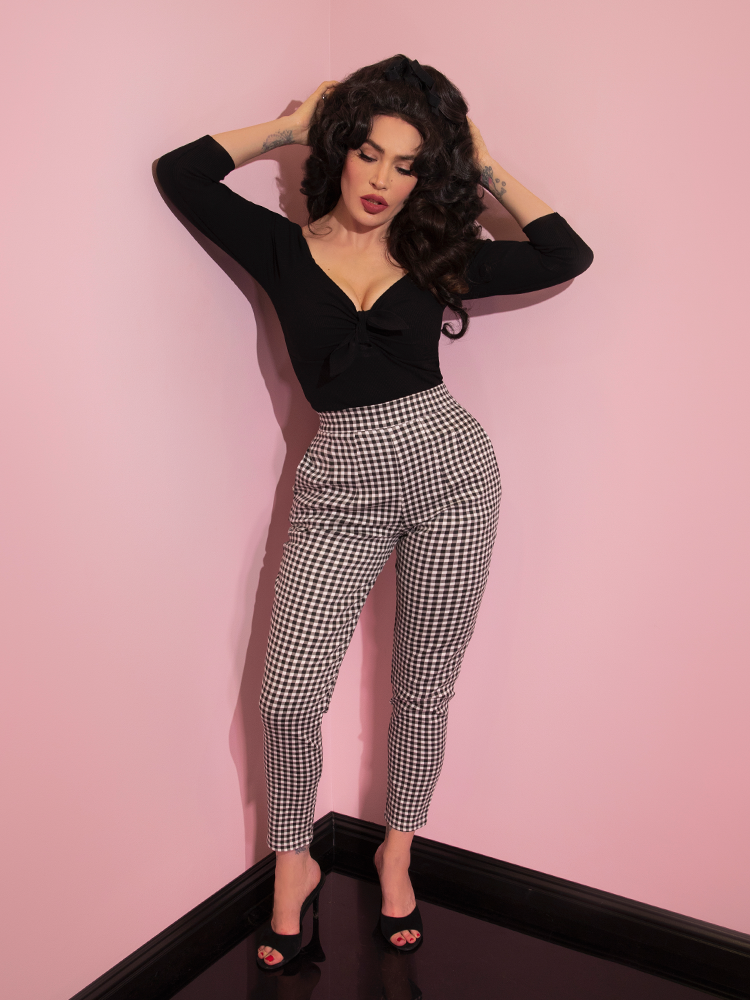Micheline Pitt running her hands through her hair while wearing a black low-cut top and black gingham cigarette pants.