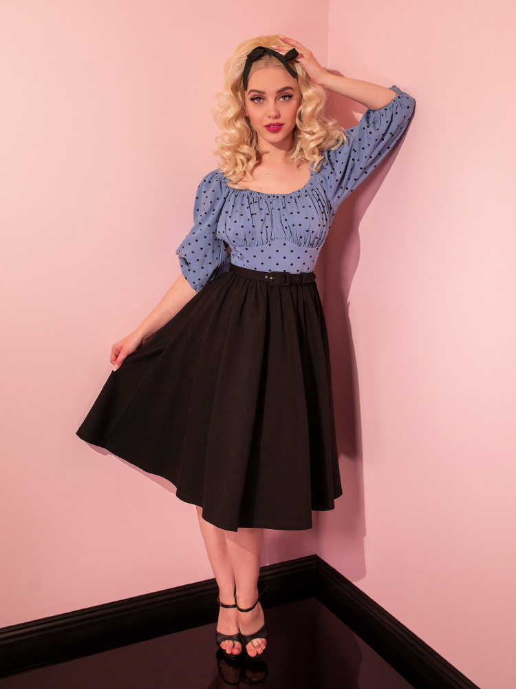 Sofia poses with one elbow against the wall and the other hand pulling out the skirt section on the Daydream Swing Dress in Sunset Blue Polka Dot.