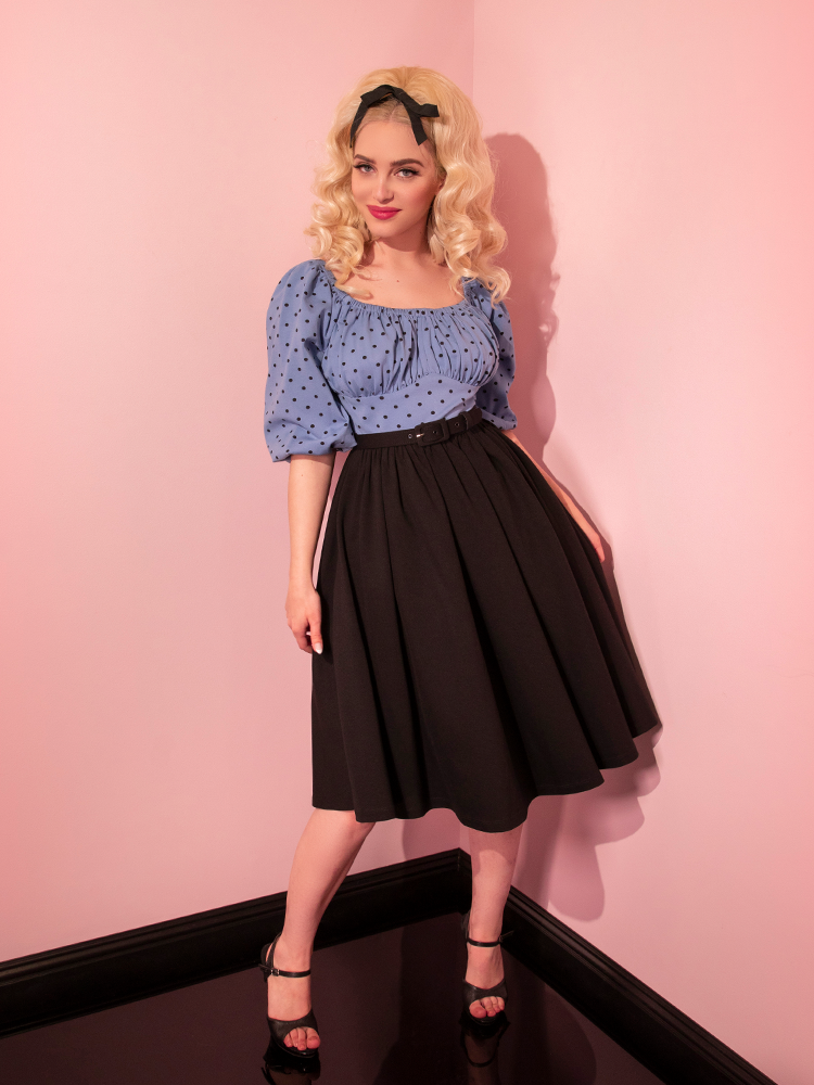 Sofia looks at the camera with her arms hanging down by the black skirt section of the Daydream Swing Dress in Sunset Blue Polka Dot.