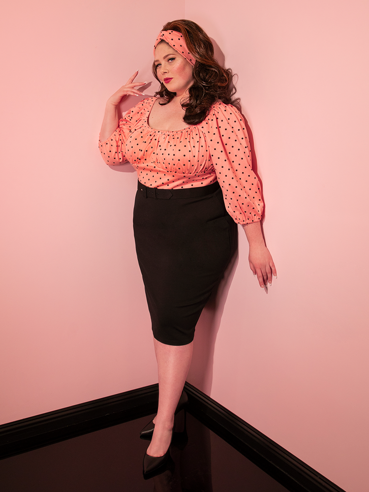 Full length shot of model posing in the corner of the room wearing a black pencil skirt paired with the Daydream Wiggle Dress in Peach Pink Polka Dot.
