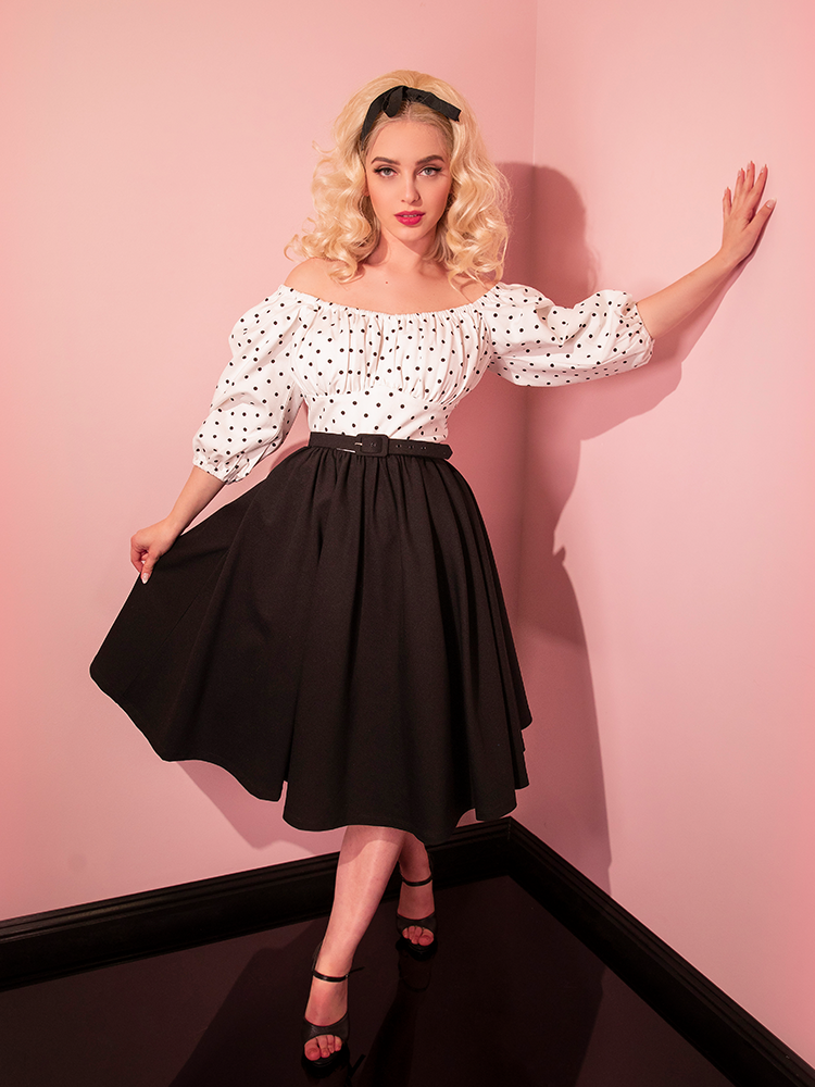 Sofia poses in a curtsy like pose while wearing the Daydream Swing Dress in Black Polka Dot.