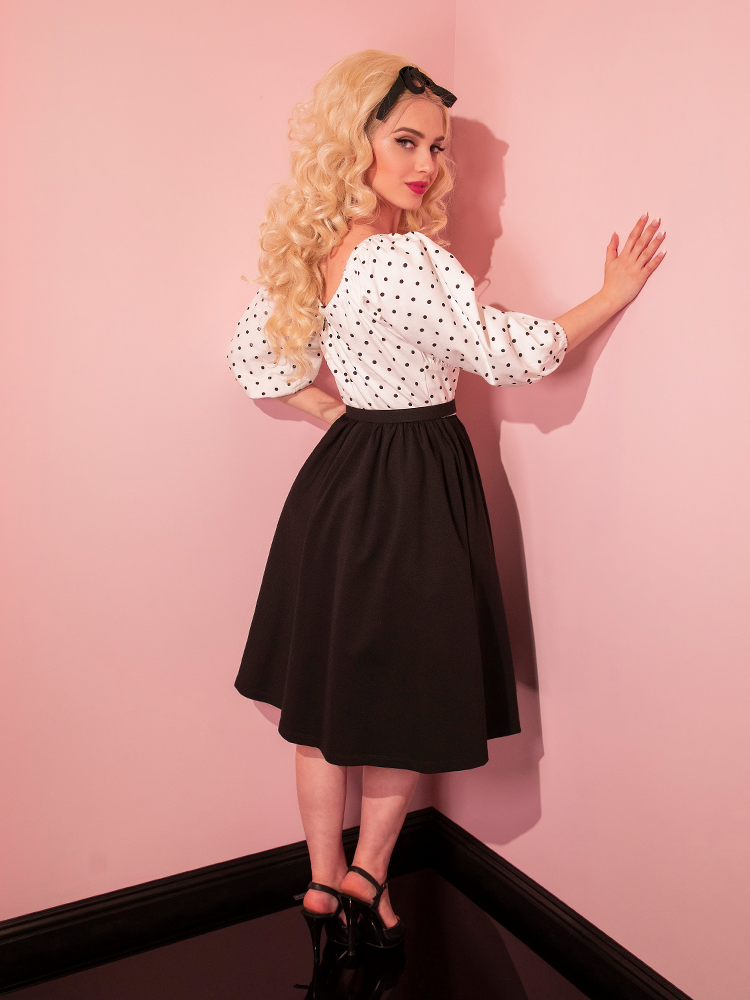 Sofia standing in the corner of her pink room wearing the Daydream Swing Dress in Black Polka Dot.