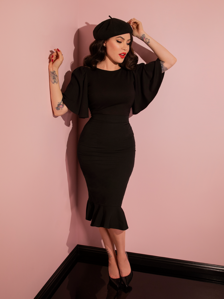 A full length shot of Micheline Pitt wearing the Deadly Kiss Top in Black tucked into a form-fitting skirt.