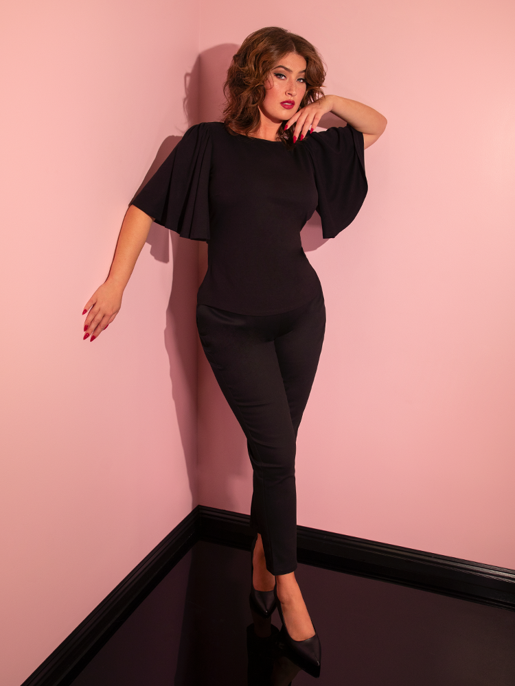Francesca posing in the corner of her pink showroom while wearing the Deadly Kiss Top in Black from retro clothing company Vixen Clothing.