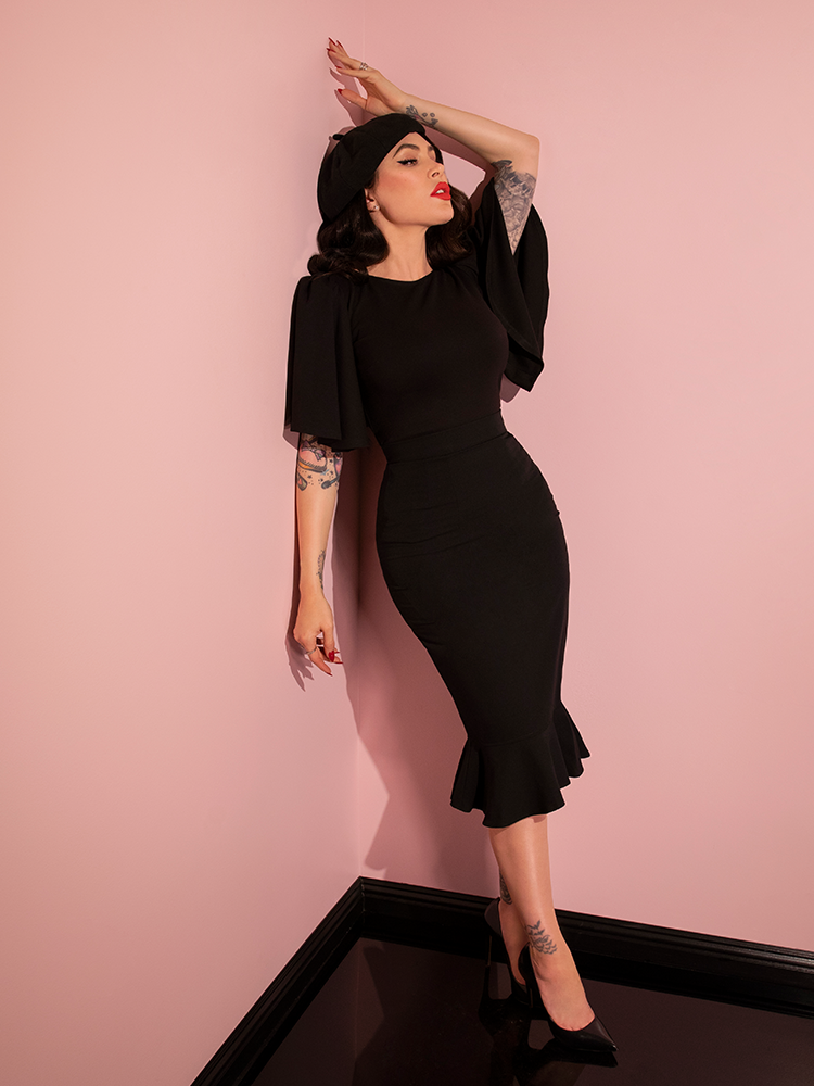 Full length image of Micheline Pitt leaning back on the pink wall of her showroom while wearing the all new retro Deadly Kiss Top in Black from Vixen Clothing.