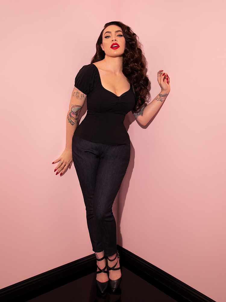 Micheline Pitt posing in the corner of her pink showroom wearing a classic retro style clothing outfit highlighted by the Cigarette Pants in Denim.