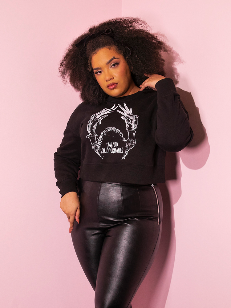 Female model wears the EDWARD SCISSORHANDS Graphic Cropped Sweatshirt in Black that falls just above the waist on her vegan leather pants.