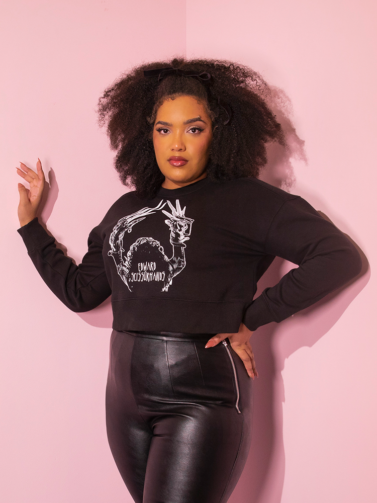 Model posing with one hand on her hip and another resting on the wall next to her while wearing the EDWARD SCISSORHANDS Graphic Cropped Sweatshirt in Black from Vixen Clothing.