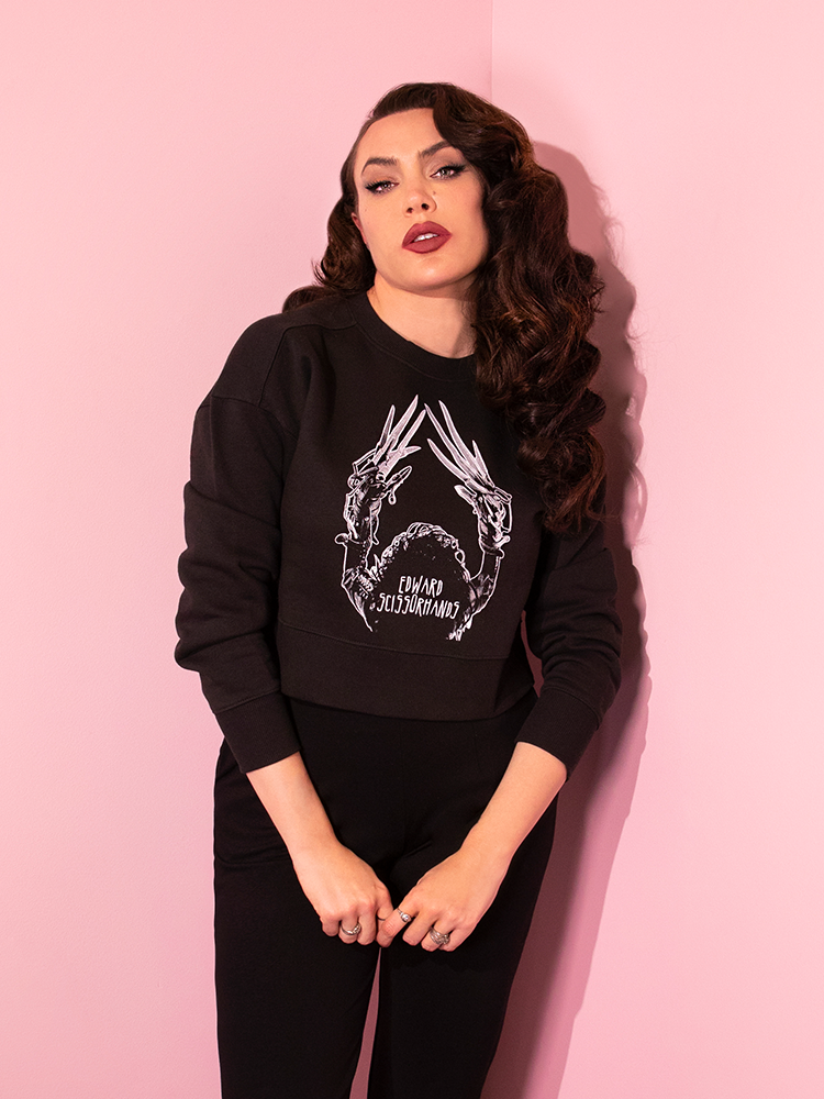 Standing with her hands clasped in front of her, Micheline Pitt models a retro style outfit accented by the EDWARD SCISSORHANDS Graphic Cropped Sweatshirt in Black.
