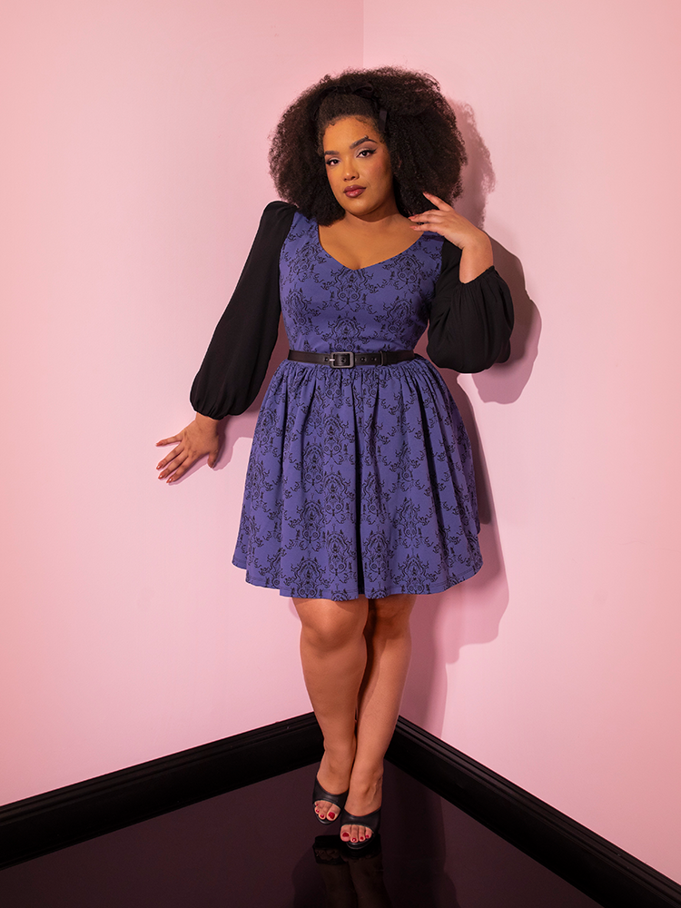Model posing with on hand help up to her shoulder, the other bracing against the pink wall behind her while wearing a retro style outfit highlighted by the EDWARD SCISSORHANDS Farewell Babydoll Dress in Wallpaper Novelty Print.