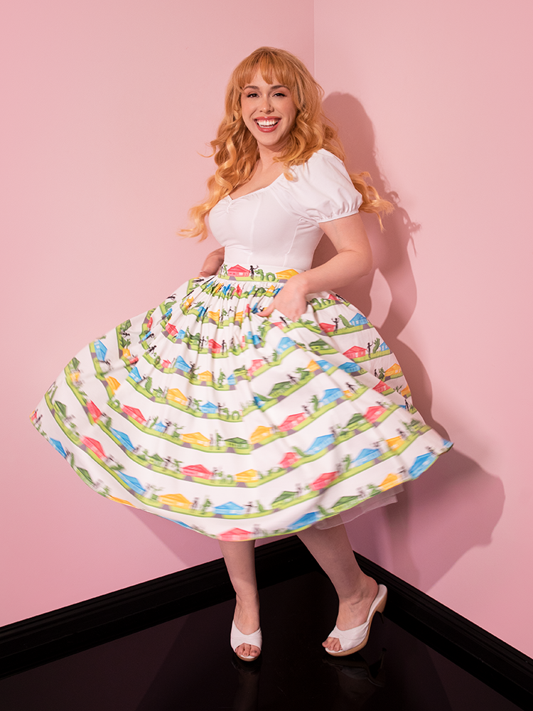 Female model smiling broadly and swinging around in the EDWARD SCISSORHANDS Swing Skirt in Suburban Topiary Novelty Print with her hands tucked into her pockets.