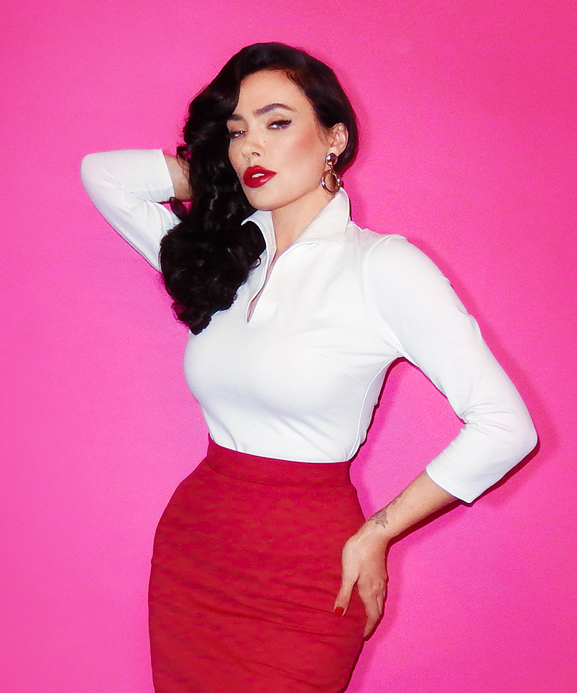 Posing with one hand on her hip and another behind her head, Micheline Pitt models a deep red pencil skirt and the Vixen Top in White.