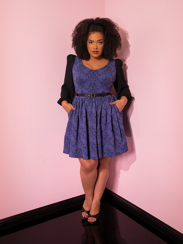 Model with her hands tucked into the pockets of the EDWARD SCISSORHANDS Farewell Babydoll Dress in Wallpaper Novelty Print from Vixen Clothing.