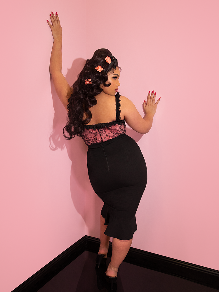 Ashleeta looking over her shoulder with pink flowers in her hair modeling the Vixen flutter skirt in black paired with a peach and black lace bustier top.