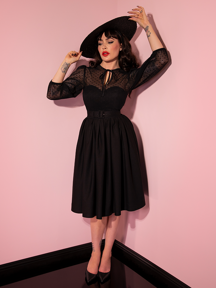 Micheline Pitt adjust her black sunhat while also wearing the Frenchie Swing Dress in Black from Vixen Clothing.