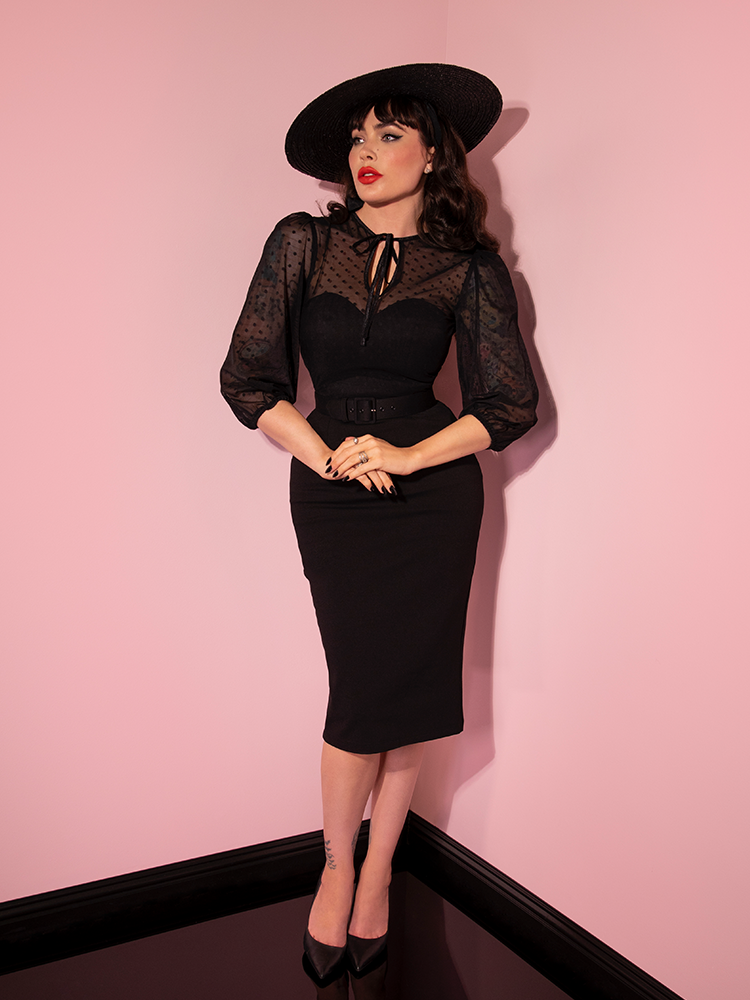 Micheline Pitt standing with her hands held at her waist while modeling the retro style dress entitled the Frenchie Wiggle Dress in Black from Vixen Clothing