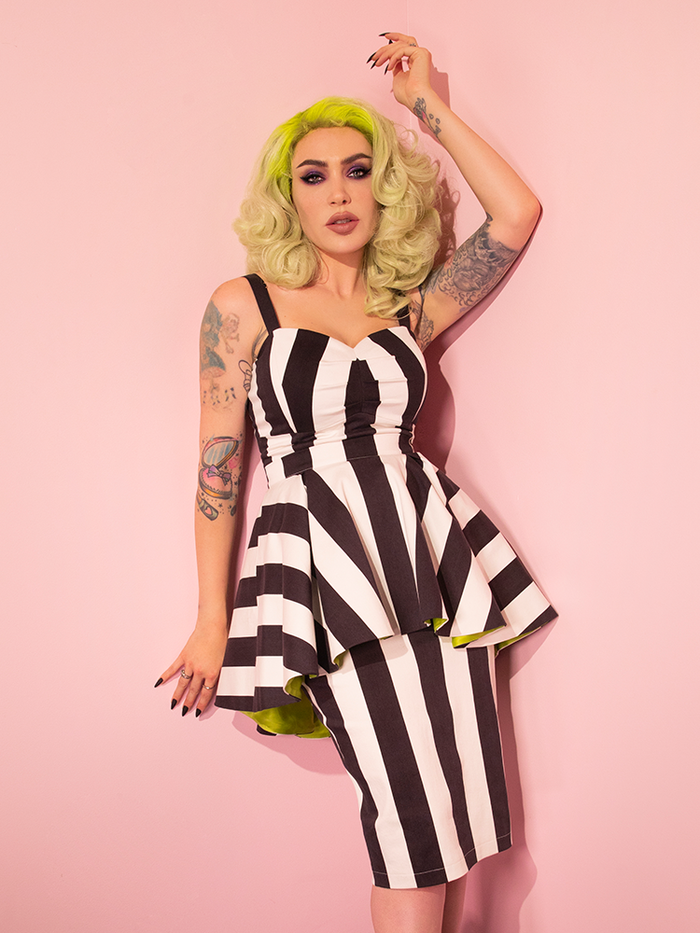 Micheline Pitt leaning against a pink wall while modeling the BEETLEJUICE™ Ghost with the Most Peplum Dress.