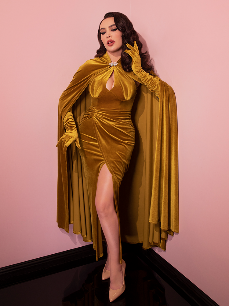 Micheline is posing in a pink room while wearing the Golden Era gown and glove set and matching cape.