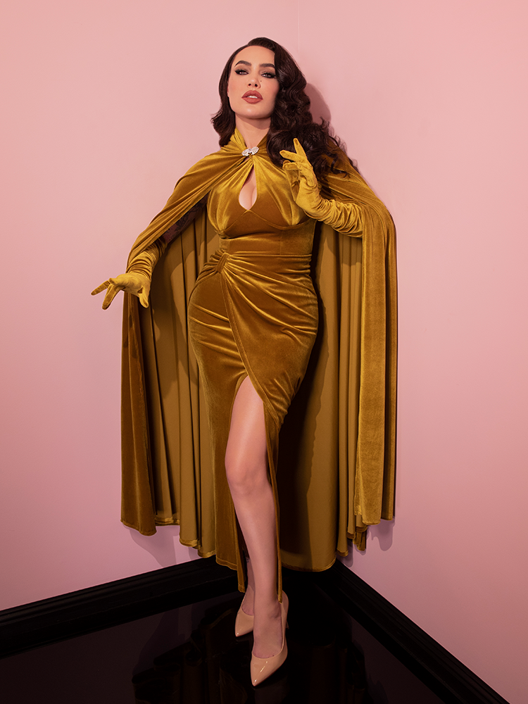 Micheline Pitt posing with her arms in positioned in front of her to show off the Golden Era Cape in Gold Velvet with matching retro inspired dress and elbow length gloves.